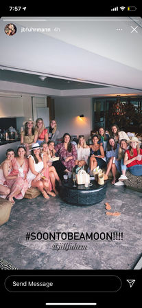 Large group of women in a bright modern lounge, wearing summer dresses and hats, smiling and holding drinks around a low coffee table with straw bags — fun bachelorette-style celebration indoors.