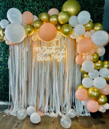 Festive peach, gold and white balloon arch with cascading ribbon-fringe photo backdrop and neon 'Til Death Do Us Party' sign on a greenery wall.