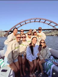 Group of smiling women on a boat wearing matching beige sweatshirts, sunglasses and shorts, posing under a rust-colored arched bridge over a calm river on a sunny day; one holds a playful face cutout while others hold drinks and beach towels.
