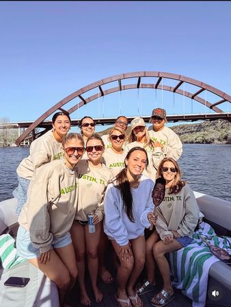 Group of smiling women on a boat wearing matching beige sweatshirts, sunglasses and shorts, posing under a rust-colored arched bridge over a calm river on a sunny day; one holds a playful face cutout while others hold drinks and beach towels.