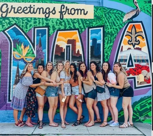 Group of women in summer outfits hugging and smiling in front of a colorful 'Greetings from NOLA' mural featuring a Mardi Gras mask, fleur-de-lis and city skyline