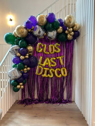 Disco-themed indoor party backdrop under a staircase with purple metallic fringe, gold foil letter balloons reading "GLOS LAST DISCO", clustered purple, green and metallic gold balloons and mirrored disco-ball decorations.