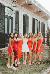 Bride in a short white dress with six bridesmaids in red and orange dresses smiling on a brick sidewalk outside a pink historic New Orleans townhouse with black shutters and decorative trim