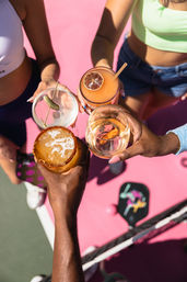 Top-down view of four hands toasting colorful summer cocktails — spiced-rim margarita, martini with a pickle, frozen citrus drink with dried lime, and a glass of white wine — over a bright pink outdoor pickleball/tennis court with a paddle visible.