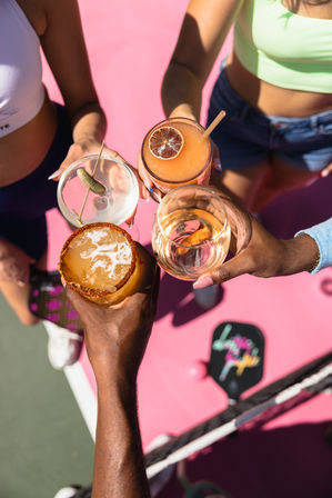 Top-down view of four hands toasting colorful summer cocktails — spiced-rim margarita, martini with a pickle, frozen citrus drink with dried lime, and a glass of white wine — over a bright pink outdoor pickleball/tennis court with a paddle visible.