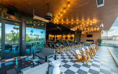 Vibrant rooftop patio dining area with checkered tile floor, string lights, wicker sofas, wooden Adirondack chairs, glass railing, palm trees and distant mountain skyline at sunset.