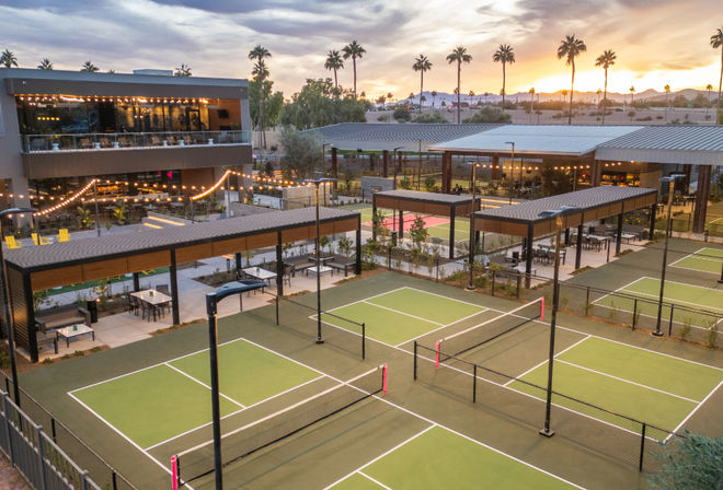 Modern outdoor pickleball courts with covered seating, string lights and palm trees silhouetted against a desert sunset and distant hills.