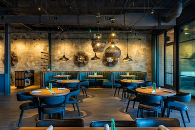Modern restaurant interior with stone accent wall, teal curved booth seating, round wooden tables set with rolled napkins, woven and metal pendant lights, exposed industrial ceiling and floor-to-ceiling glass entrance — warm contemporary dining space.