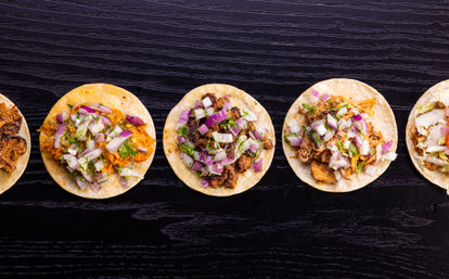 Row of colorful Mexican street tacos on corn tortillas topped with shredded and grilled meats, diced red onion, and cilantro on a dark wooden table.