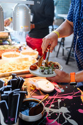 Hand placing sushi rolls, a glazed bun and a taco onto a paper plate at a colorful event buffet food station with tongs, dipping sauces, chips and stacked napkins visible.