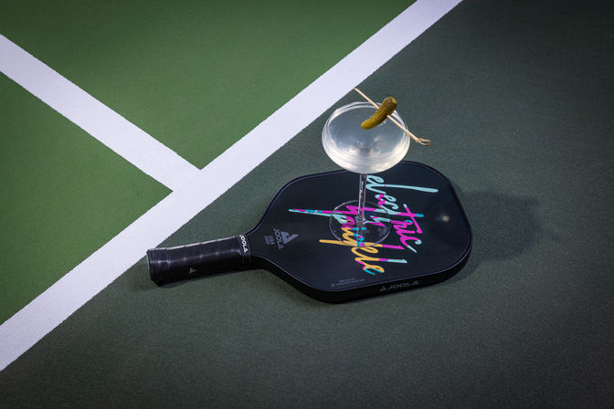 Pickleball paddle on a green hardcourt beside white boundary lines, topped by a coupe glass holding a clear cocktail garnished with a small pickle — playful sports-and-drinks still life.