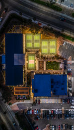 Aerial night view of illuminated tennis courts and an adjoining sports complex with glowing roofs, outdoor seating, parked cars, palm trees and surrounding city streets.