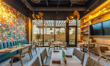 Contemporary restaurant interior with a vibrant floral mural, teal banquette seating, wooden tables and woven chairs, rattan pendant lights, and glass doors opening to an outdoor patio at sunset.