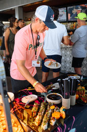 Attendee at an outdoor food festival picking sushi rolls from a colorful buffet table with soy dipping sauce, disposable plates, chopsticks, and a casual pop-up bar in the background.