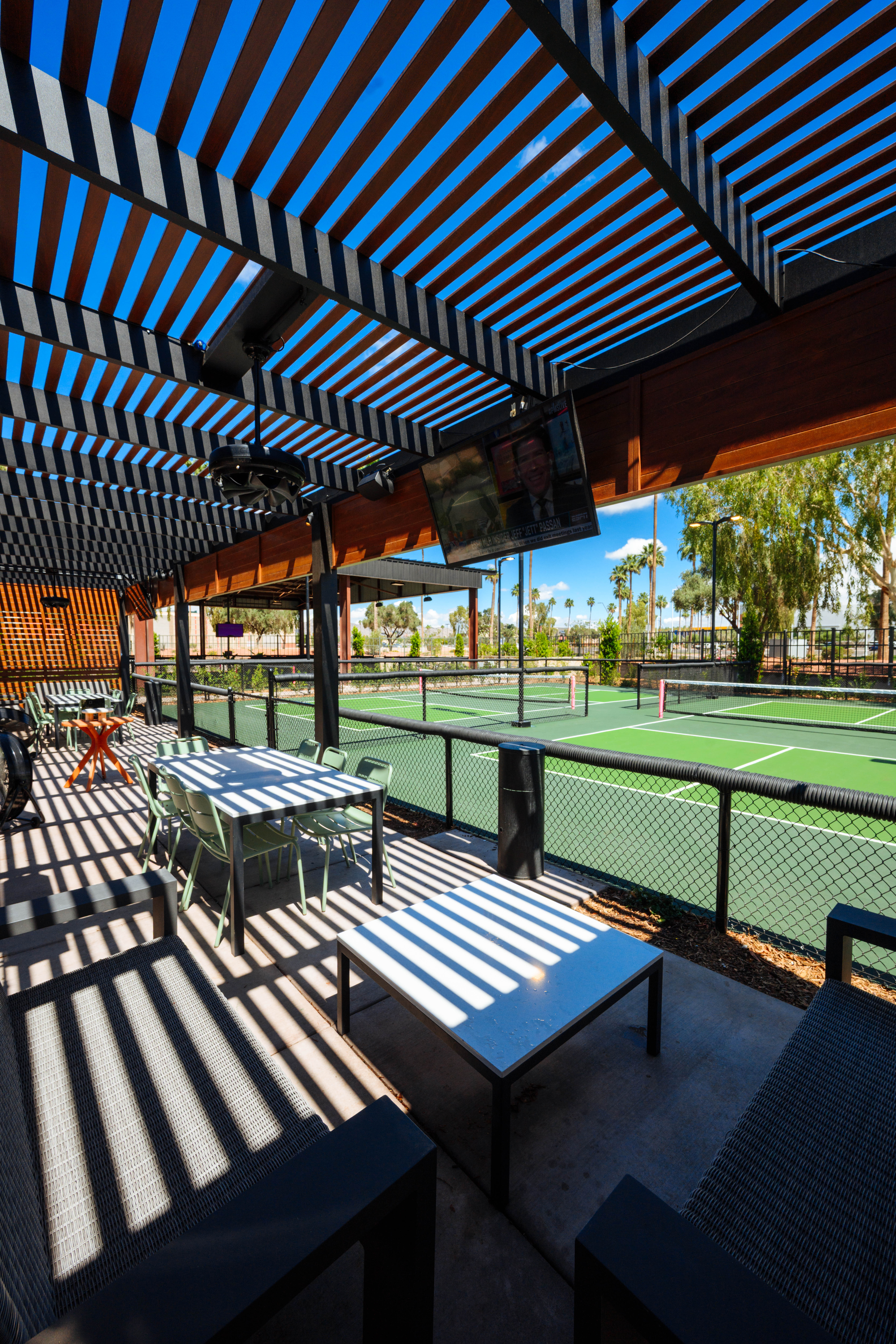 Shaded pergola patio casting striped shadows over tables and lounge seating, overlooking bright green outdoor pickleball courts, palm trees and clear blue sky.