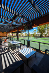 Shaded pergola patio casting striped shadows over tables and lounge seating, overlooking bright green outdoor pickleball courts, palm trees and clear blue sky.