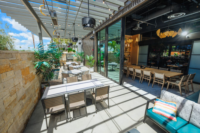 Sun-drenched outdoor patio dining area with a slatted pergola, glass sliding doors to a modern indoor dining room, wooden tables and chairs, potted tropical plants, string lights and a cushioned lounge with colorful pillow.