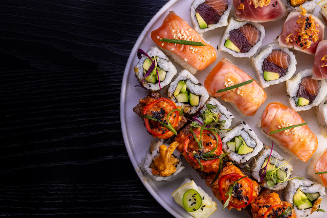 Overhead shot of an assorted sushi platter — salmon nigiri, avocado maki, spicy tuna and specialty rolls garnished with sesame seeds and microgreens on a white plate against a dark wood table.