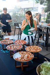 Woman choosing slices from a pizza buffet on an outdoor patio — several thin‑crust pies on raised stands at a casual food event.