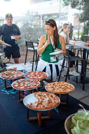 Woman choosing slices from a pizza buffet on an outdoor patio — several thin‑crust pies on raised stands at a casual food event.