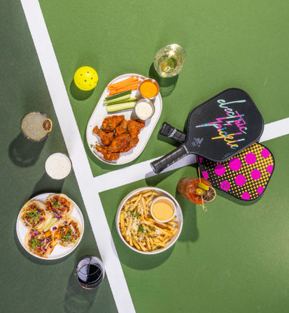 Top-down view on a green pickleball court with paddles and yellow ball beside plates of chicken wings with dips, fries, tacos, veggie sticks and assorted drinks — court-side snacks