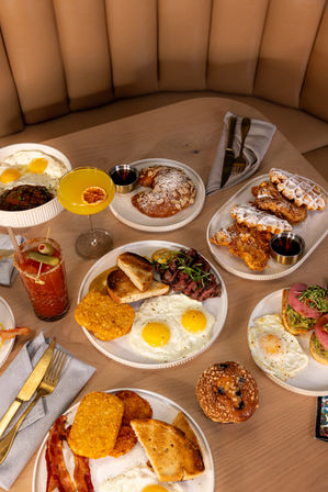 Colorful brunch spread on a wooden table in a cozy booth: plates of sunny-side eggs with hash browns, bacon, toast and steak, almond croissant, chicken-and-waffles, muffin, mimosa and Bloody Mary cocktail.