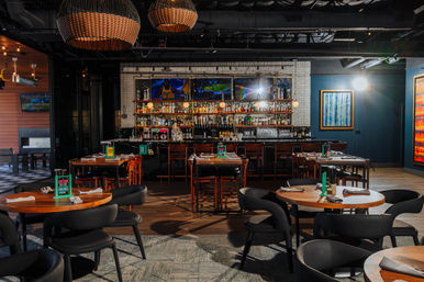 Modern bar and restaurant interior with round wooden tables and black chairs, woven pendant lights, a stocked back bar with TVs, and colorful framed artwork