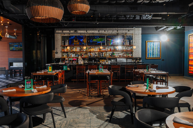 Modern bar and restaurant interior with round wooden tables and black chairs, woven pendant lights, a stocked back bar with TVs, and colorful framed artwork