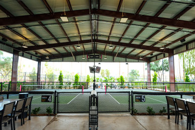 Covered outdoor community courts under a metal pavilion, fenced and numbered 4 and 5 with nets, patio seating and landscaped palms