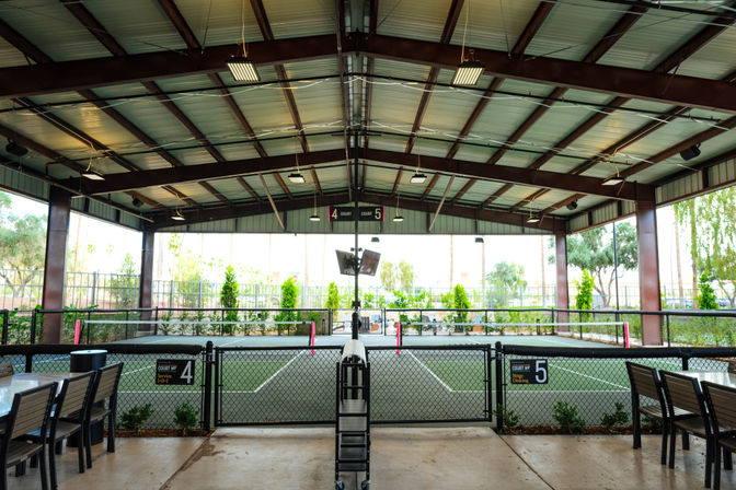 Covered outdoor community courts under a metal pavilion, fenced and numbered 4 and 5 with nets, patio seating and landscaped palms