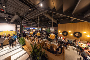 Bustling modern indoor food hall and restaurant with wooden booths, round straw pendant lights, industrial open ceiling, a lively bar with TV screens in the background, and groups dining together.