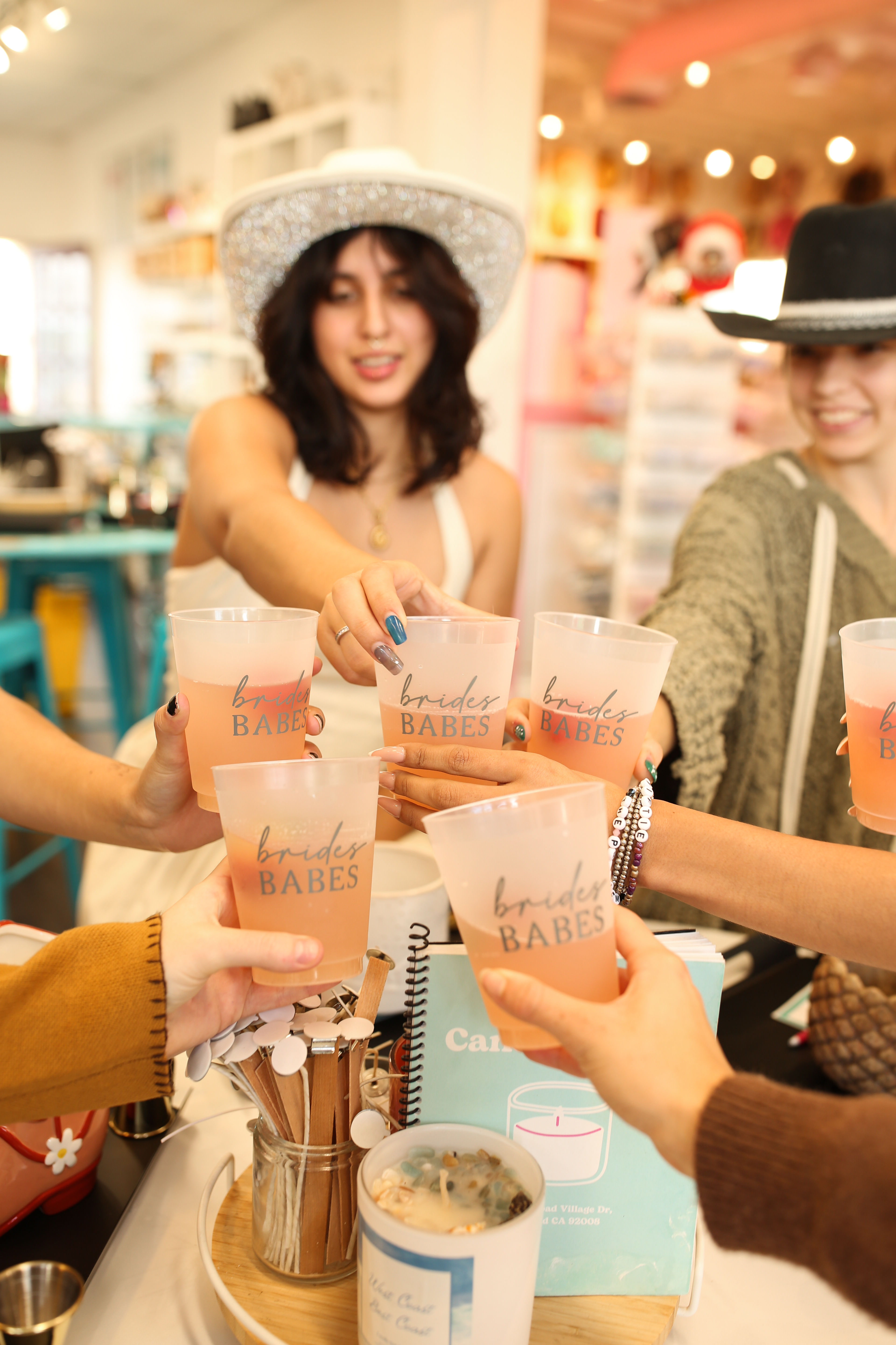 Bachelorette party toast in a colorful boutique — friends clinking 'Brides Babes' cups filled with pink drinks