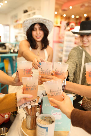 Bachelorette party toast in a colorful boutique — friends clinking 'Brides Babes' cups filled with pink drinks