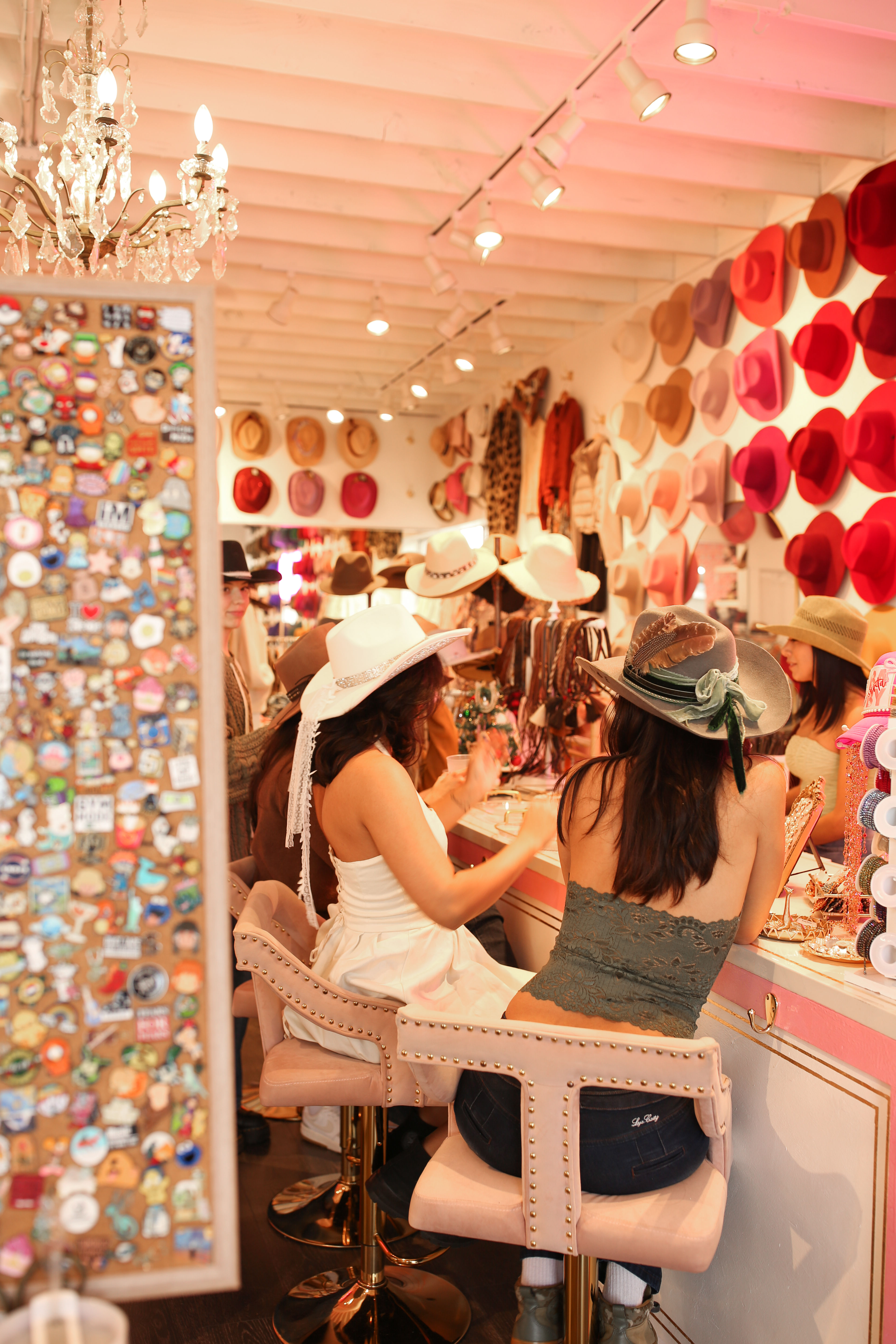 Shoppers trying on cowboy and felt hats at a trendy hat boutique with a pink-and-red hat wall, mirrored jewelry counter, chandelier lighting and studded pink bar stools