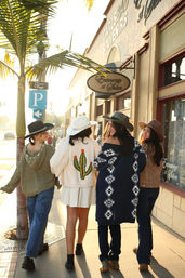 Four friends in wide‑brim hats and cozy cardigans—one with a cactus knit—walking arm‑in‑arm down a sunny California beach‑town main street with palm trees and storefronts at golden hour.