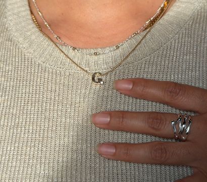 Close-up of a cream knit sweater with layered gold and silver necklaces, one featuring a small initial pendant, and a hand with natural nails wearing a sculptural silver ring resting on the chest.