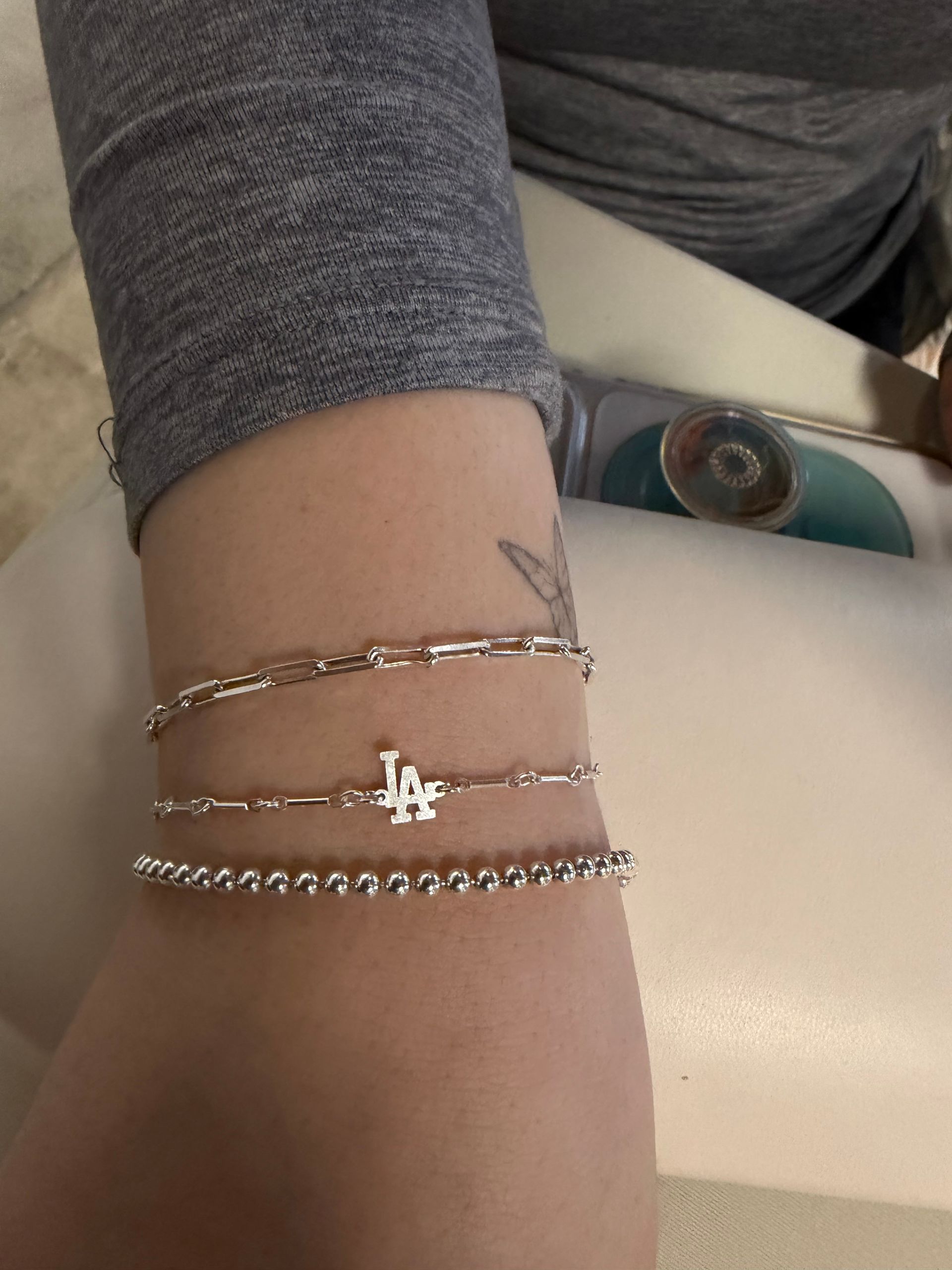 Forearm resting on a table wearing layered silver bracelets — beaded strand, thin link chains and an 'LA' charm — small tattoo peeking, gray sleeve and blue contact lens case nearby.
