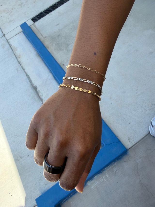 Close-up of a forearm with a stack of three delicate gold and silver chain bracelets and a black metal ring, hand resting above a concrete sidewalk next to a blue painted curb.