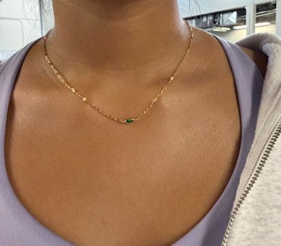 Close-up of a person's neck and collarbone wearing a delicate gold chain with a small green teardrop pendant over a lavender top and light zip-up hoodie in an indoor setting
