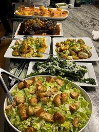 Home-style buffet on a countertop with a large crouton-topped romaine salad, cheesy kale, two plates of roasted baby potatoes with scallions, glazed braised short ribs, and lemon-topped baked salmon with green sauce.
