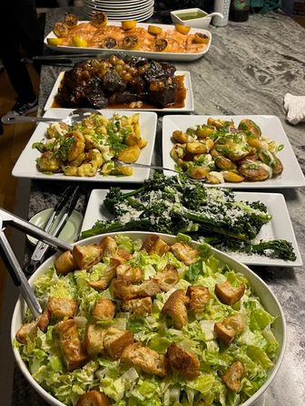 Home-style buffet on a countertop with a large crouton-topped romaine salad, cheesy kale, two plates of roasted baby potatoes with scallions, glazed braised short ribs, and lemon-topped baked salmon with green sauce.