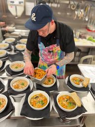 Chef in a busy commercial kitchen wearing a colorful apron and navy cap plating bowls of hearty bean and vegetable soup with crispy garnish and a slice of bread.
