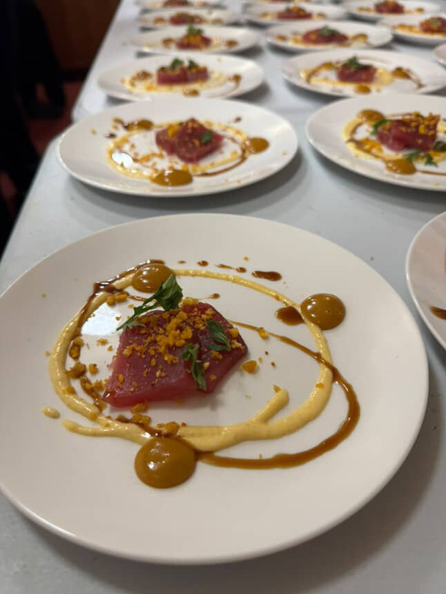 Plated tuna crudo appetizer — bright pink tuna topped with microgreens and crunchy crumbs, playful mustard and brown sauce swirls on white plates lined on a restaurant prep table.