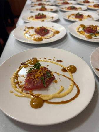 Plated tuna crudo appetizer — bright pink tuna topped with microgreens and crunchy crumbs, playful mustard and brown sauce swirls on white plates lined on a restaurant prep table.