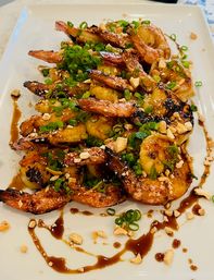 Close-up of grilled shrimp and caramelized plantains topped with chopped peanuts and scallions, drizzled with sweet soy glaze on a white platter