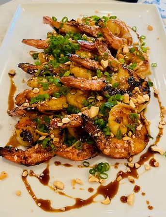 Close-up of grilled shrimp and caramelized plantains topped with chopped peanuts and scallions, drizzled with sweet soy glaze on a white platter