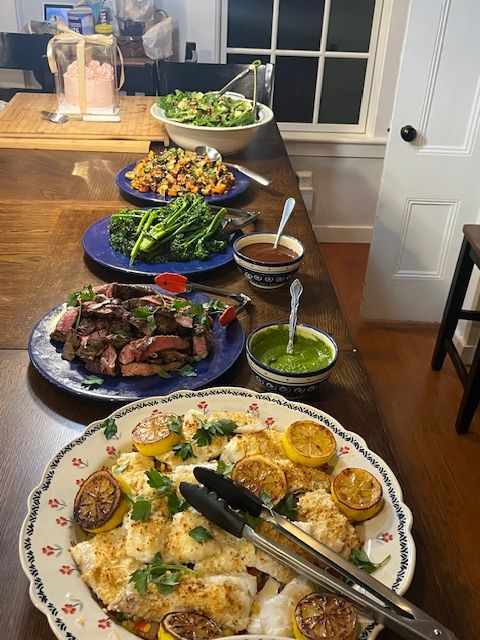 Home kitchen counter with family-style dinner spread: platter of baked white fish topped with charred lemon slices and herbs, sliced grilled steak, broccolini, roasted squash and sweet-potato salad, mixed greens and dipping sauces.