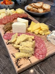 Charcuterie board on a wooden serving board with assorted cheeses, sliced salami, whole grain mustard and fruit jam, plus a bowl of toasted crostini and pickled vegetables in the background.