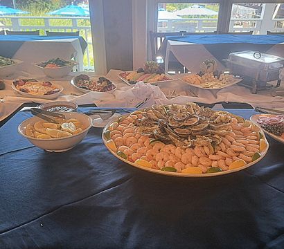 Seafood buffet: large platter of peeled shrimp and oysters arranged with lemon wedges and cocktail sauce on a navy tablecloth, indoor spread near sunlit windows overlooking outdoor patio umbrellas.