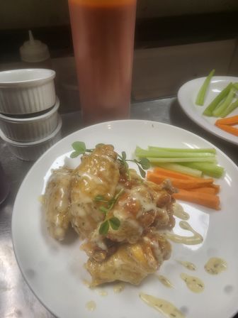 Sticky glazed chicken wings with creamy herb drizzle and microgreens, served with celery and carrot sticks on a white plate in a commercial kitchen.